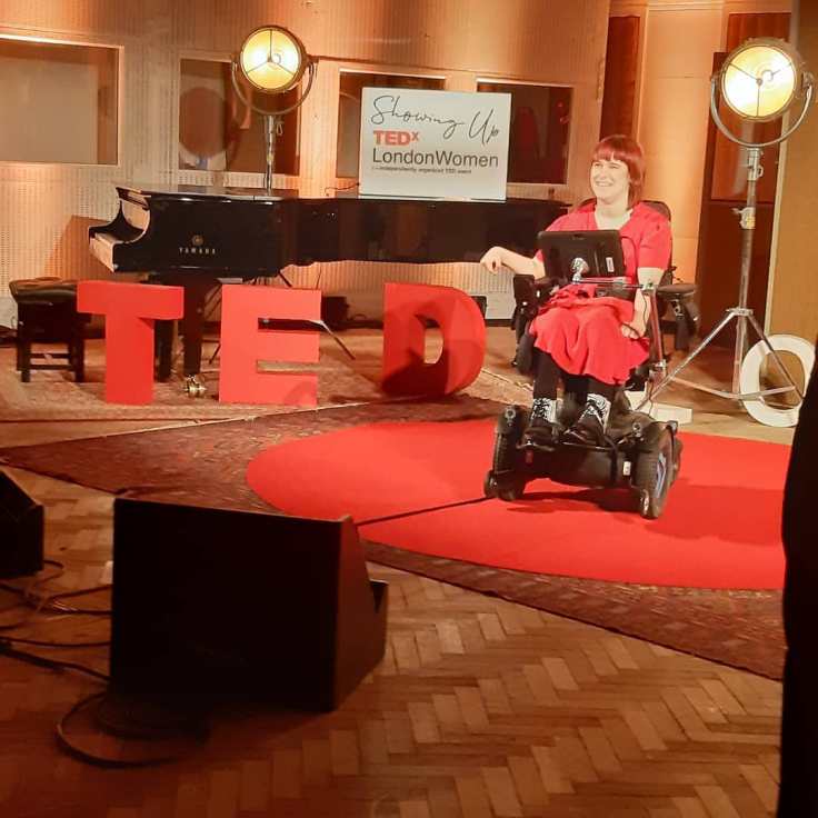 In this photo Clair Malone (she/her), physicist, member of the LGBTQ+ community, white disabled woman, is smiling, wearing a red dress, black leggings, and zebra print shoes, with shoulder length red hair, in her wheelchair, in the centre of a TED X red circle carpet, in front of a large TED X sign, with a grand piano in the background. On the piano, a white sign reads: Showing Up TED X London